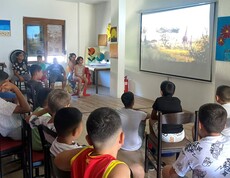 Mehrere Kinder sitzen in einem Raum auf Stühlen und schauen gemeinsam auf eine große Leinwand, auf der ein Film mit Tieren in einer Landschaft läuft.