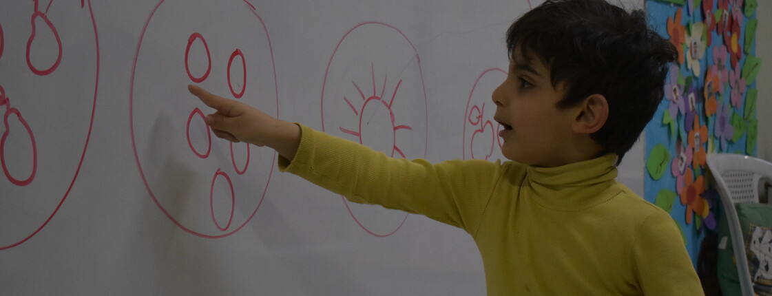 Kind beim Malen und Lernen in der Kinderkrippe Damaskus Ein Kind in der Kinderkrippe Damaskus zeigt auf eine Zeichnung an der Wand, während es etwas erklärt. Es ist mit einem gelben Pullover gekleidet und schaut konzentriert.