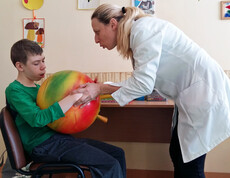 Eine Therapeutin unterstützt ein Kind im Kinderdorf Gomel bei einer medizinischen Übung mit einem großen Ball, um seine Motorik und Beweglichkeit zu fördern.