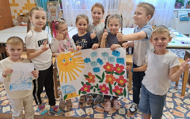 Mehrere Kinder stehen in einem Raum und halten selbst gestaltete bunte Bilder und Collagen hoch.