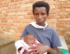 Eine Mutter in Burundi hält ihr Baby in den Armen, sitzt vor einer Ziegelwand und blickt ernst in die Kamera.