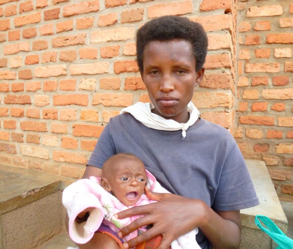 Eine Mutter in Burundi hält ihr Baby in den Armen, sitzt vor einer Ziegelwand und blickt ernst in die Kamera.