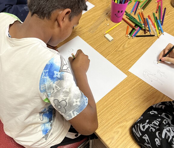 Ein Kind sitzt an einem Tisch und zeichnet mit einem Buntstift auf ein Blatt Papier, während weitere Kinder daneben ebenfalls malen; auf dem Tisch liegen viele bunte Stifte.
