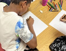 Ein Kind sitzt an einem Tisch und zeichnet mit einem Buntstift auf ein Blatt Papier, während weitere Kinder daneben ebenfalls malen; auf dem Tisch liegen viele bunte Stifte.