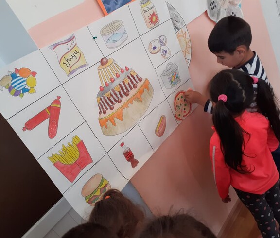 Zwei Kindergartenkinder stehen vor einem selbstgemalten Plakat, dass ungesunde Lebensmittel zeigt.