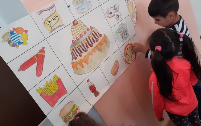 Zwei Kindergartenkinder stehen vor einem selbstgemalten Plakat, dass ungesunde Lebensmittel zeigt.