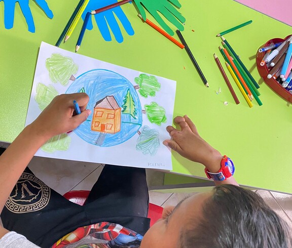 Ein Kind malt ein farbiges Bild mit einem Haus, Bäumen und blauem Himmel; auf dem Tisch liegen viele Buntstifte und bunte Papierhände.