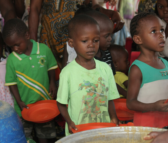 Mehrere Kinder stehen in einer Schlange mit leeren orangefarbenen Tellern in der Hand und warten auf eine Essensausgabe. Im Vordergrund ist ein großer Topf mit Brei oder Suppe zu sehen, aus dem gerade geschöpft wird. Die Kinder blicken ernst oder geduldig nach vorne, im Hintergrund sind weitere Kinder und Erwachsene zu erkennen.