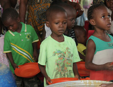 Mehrere Kinder stehen in einer Schlange mit leeren orangefarbenen Tellern in der Hand und warten auf eine Essensausgabe. Im Vordergrund ist ein großer Topf mit Brei oder Suppe zu sehen, aus dem gerade geschöpft wird. Die Kinder blicken ernst oder geduldig nach vorne, im Hintergrund sind weitere Kinder und Erwachsene zu erkennen.