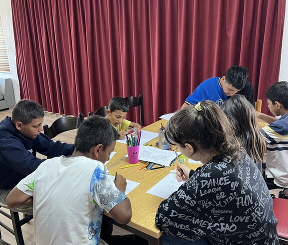 Mehrere Kinder sitzen um einen Tisch und malen mit Buntstiften auf Papier; auf dem Tisch stehen Becher mit Stiften.