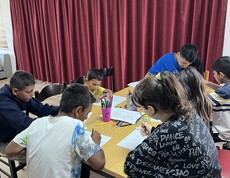 Mehrere Kinder sitzen um einen Tisch und malen mit Buntstiften auf Papier; auf dem Tisch stehen Becher mit Stiften.