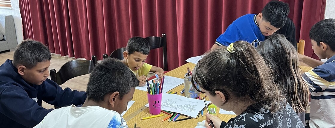 Mehrere Kinder sitzen um einen Tisch und malen mit Buntstiften auf Papier; auf dem Tisch stehen Becher mit Stiften.