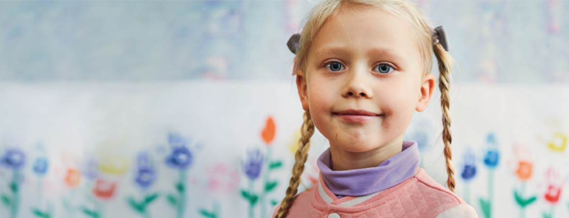 Patenschaften für Kinder weltweit Ein junges Mädchen mit blonden Zöpfen steht vor einer bunten Wand mit gemalten Blumen und lächelt freundlich