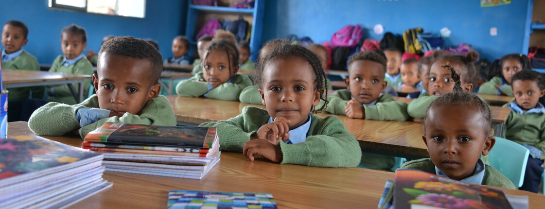 mehrere Kinder sitzen in einem Klassenzimmer und schauen von ihren Schulbänken in die Kamera