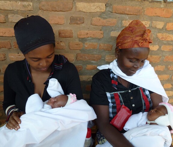 Zwei Frauen in Burundi halten ihre Neugeborenen liebevoll in den Armen, während sie vor einer Ziegelwand sitzen.