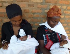 Zwei Frauen in Burundi halten ihre Neugeborenen liebevoll in den Armen, während sie vor einer Ziegelwand sitzen.