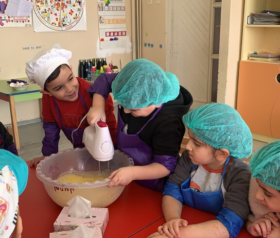 Kindergartenkinder, die verschiedene Kochmützen tragen, stehen um einen Tisch. Ein Kind rührt Teig mit einem Stabmixer.