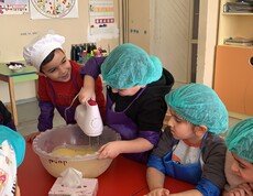 Kindergartenkinder, die verschiedene Kochmützen tragen, stehen um einen Tisch. Ein Kind rührt Teig mit einem Stabmixer.