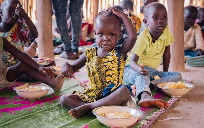 Kinder sitzen auf einer bunten Matte und essen gemeinsam in einer Hilfseinrichtung. Im Hintergrund sind weitere Kinder zu sehen