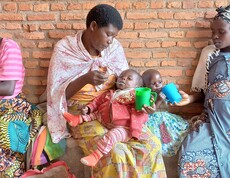 Mehrere Frauen sitzen mit kleinen Kindern auf einer Bank vor einer Backsteinwand und füttern sie.