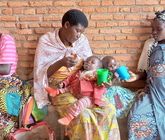 Mehrere Frauen sitzen mit kleinen Kindern auf einer Bank vor einer Backsteinwand und füttern sie.