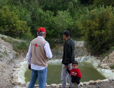 Caritas-Mitarbeiter und Landwirt mit Kind stehen vor einem neu angelegten Regenwasserspeicher in Deir Touma