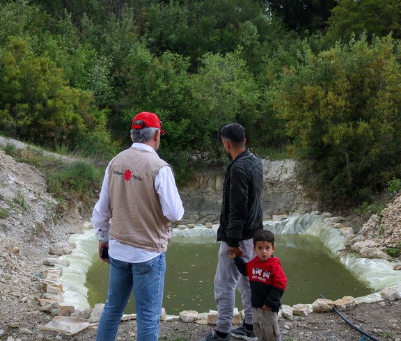 Caritas-Mitarbeiter und Landwirt mit Kind stehen vor einem neu angelegten Regenwasserspeicher in Deir Touma