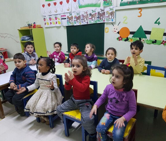 Eine Gruppe von Kindern in einer Tagesstätte in Damaskus sitzt fröhlich an einem Tisch, einige klatschen und spielen, während sie betreut werden