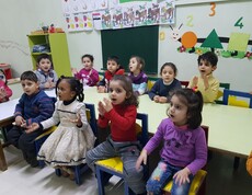 Eine Gruppe von Kindern in einer Tagesstätte in Damaskus sitzt fröhlich an einem Tisch, einige klatschen und spielen, während sie betreut werden