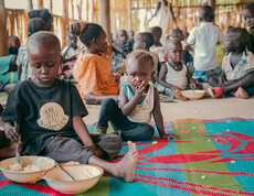 Kinder sitzen auf einer bunten Matte und essen in einem Ernährungszentrum im Südsudan.
