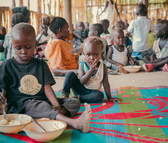 Kinder sitzen auf einer bunten Matte und essen in einem Ernährungszentrum im Südsudan.